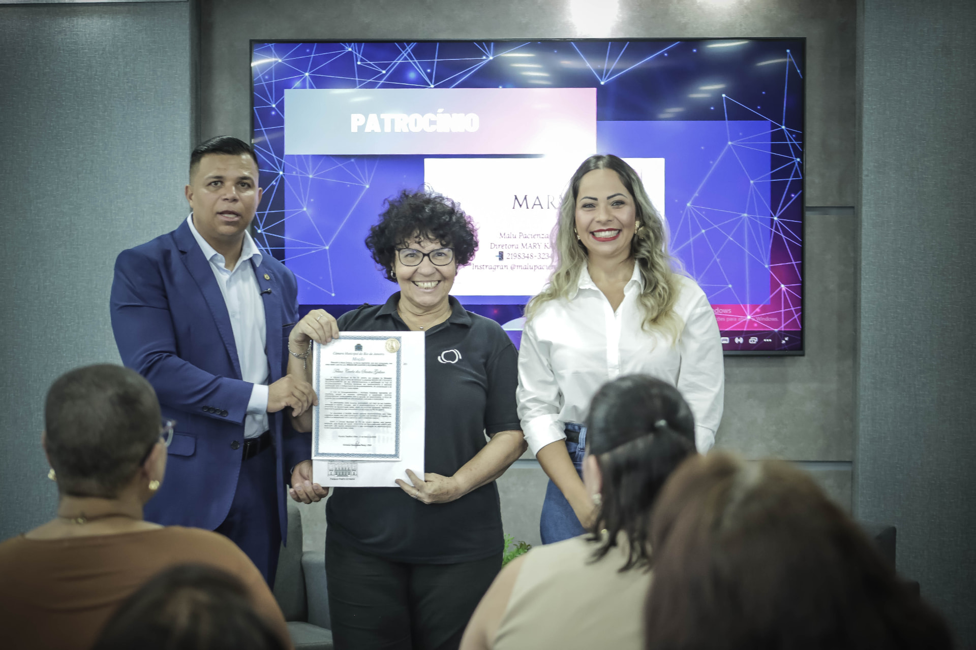 Representantes do evento Resenha de Negócios entregam uma moção de reconhecimento à Omega Foto e Vídeo, com a homenageada segurando o diploma no palco